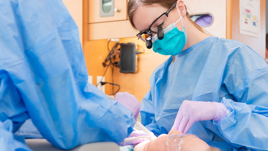 A dental student in surgical attire performs a procedure on a patient in a dental clinic.