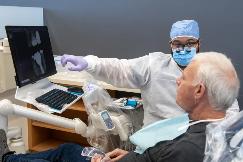 A dental provider reviews X-ray images on a monitor with a patient seated in a dental chair.