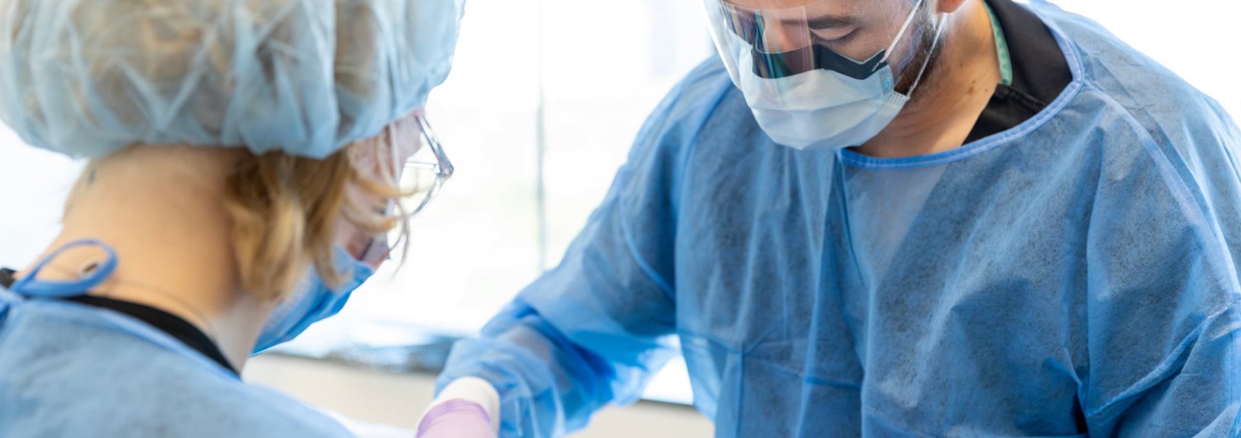 Two dental surgical professionals wearing blue scrubs, sterile caps, face masks, and protective face shields working together during an oral surgery procedure.