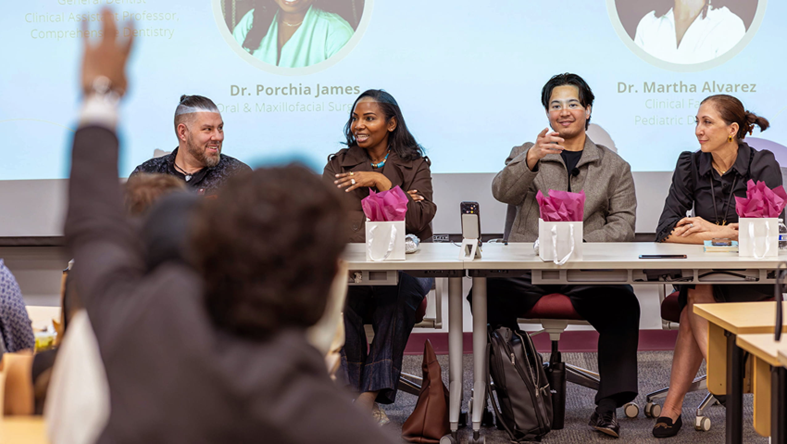 An audience member raises a hand to ask a question during an academic panel discussion featuring dental faculty members on stage.
