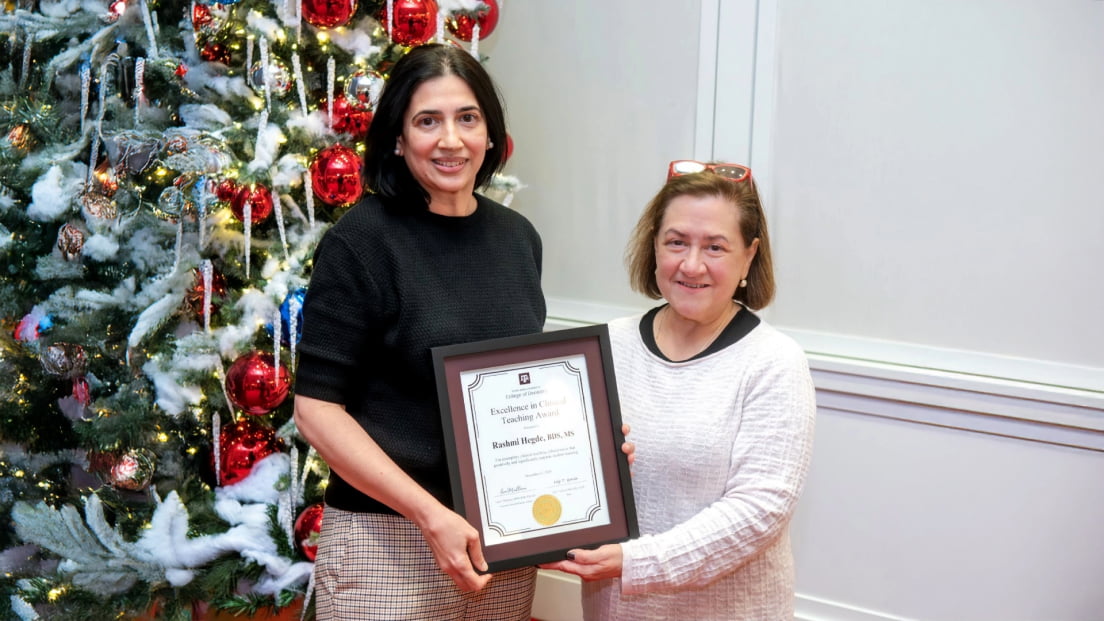 Dr. Rashmi Hegde smiling alongside a colleague while holding her framed Excellence in Clinical Teaching Award certificate in front of a decorated holiday tree.