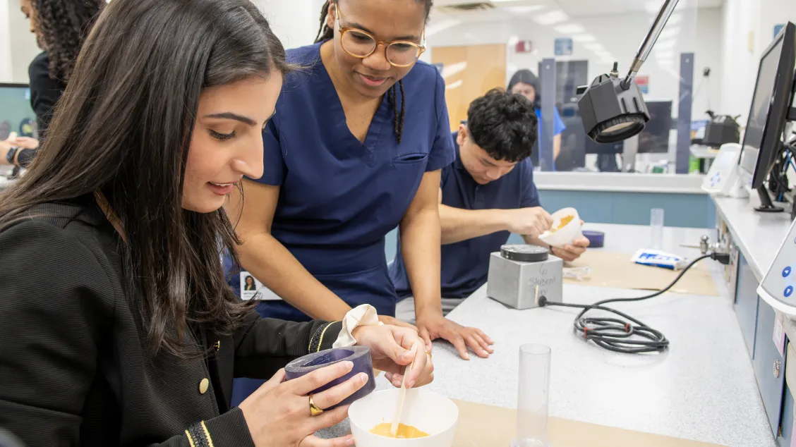 Two dental students work together on a lab exercise at a clinic workstation, with a third student visible in the background.