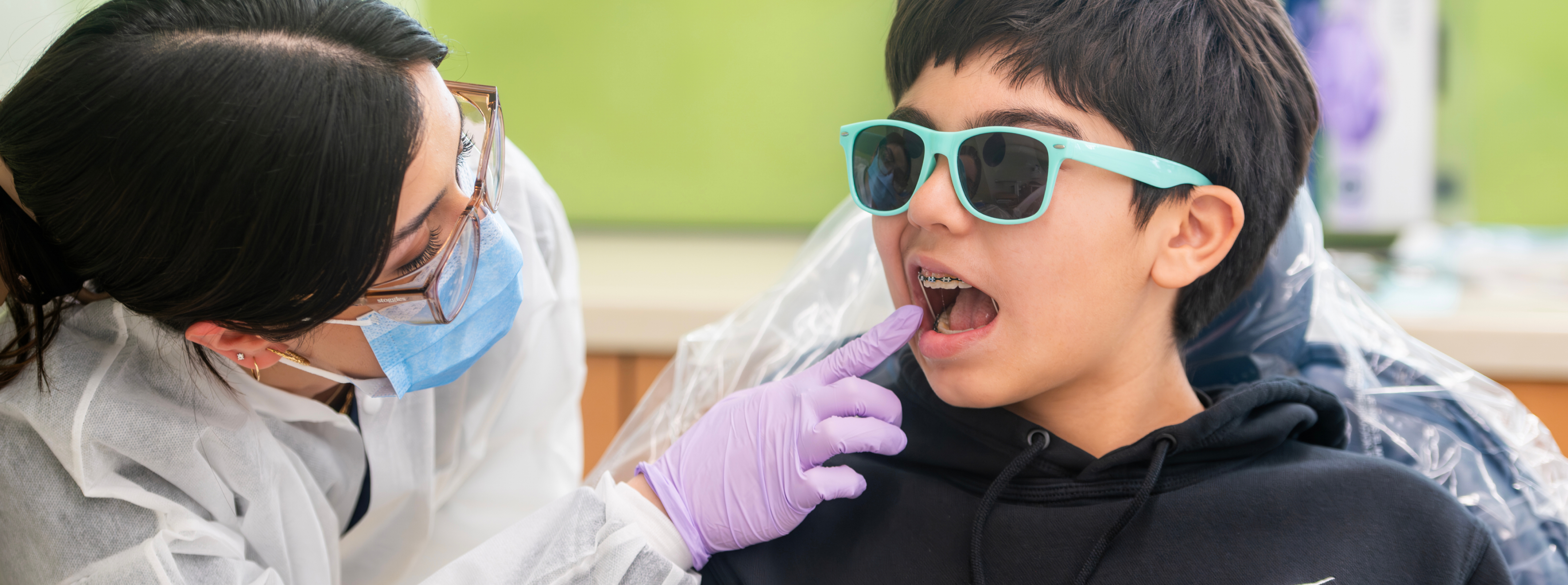 A dental student examines a young patient wearing teal protective sunglasses during a dental appointment.