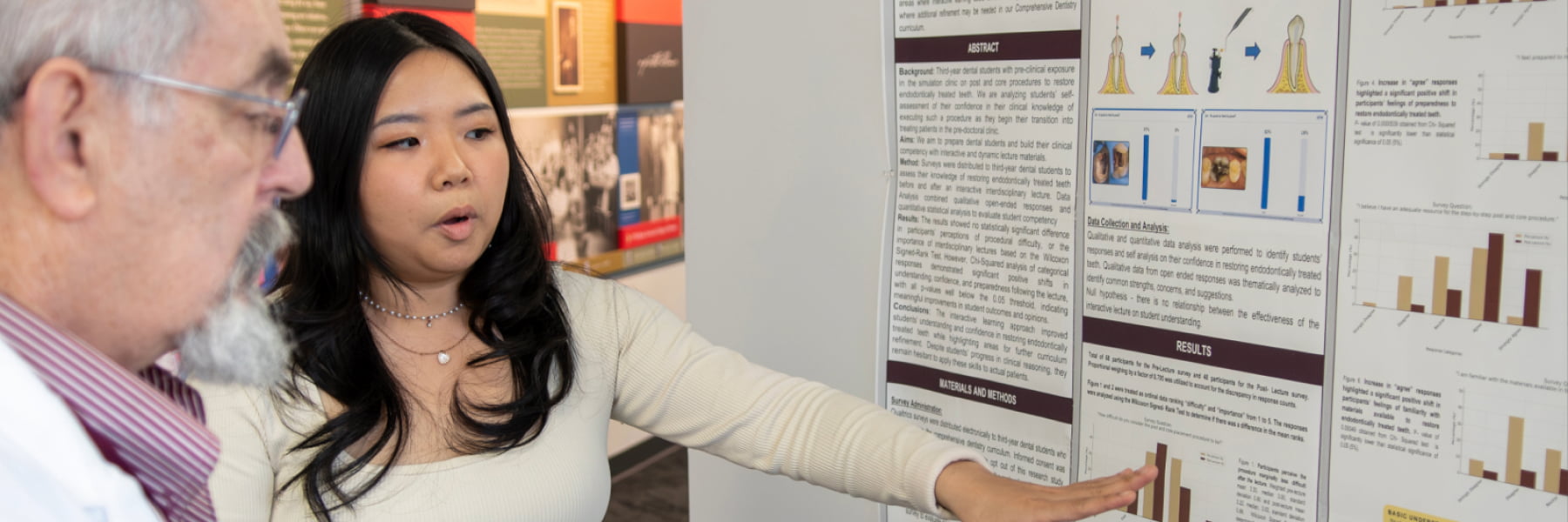 A young female student presenting her research findings to an older faculty member in front of a detailed academic research poster.